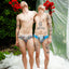 Two men standing outdoors with tattoos and colorful underwear, surrounded by flowers and white fabric.