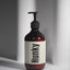 An amber glass pump bottle of Hunky Handmade Body Wash with a black pump and a cream label, set against a modern gray background with vertical ribbed panels.