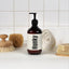 An amber glass pump bottle labeled 'Hunky' by The Ordinary, styled on a white surface with a white tiled background and bath accessories including a towel, sponge, brush, and soap bar.