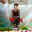 POC nonbinary person wearing black Cotton Long Tank Binder tucked into Grey Monocrome underwear peaking out under black mesh shorts over that that say Trans I am in gold letters  holding flowers in front of a table with red flowers and green plants, with a building in the background.