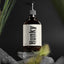 An amber glass pump bottle labeled 'Hunky' by 'TGS" Handsome Body Wash sits on stacked gray stones with blurred green grass against a dark background.