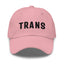 A pink dad hat with the word 'TRANS' embroidered in white on the front.